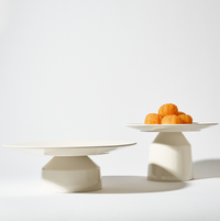Two white plates, including a Solare Serving Tray on a stand, are displayed against a plain white background; the plate on the left is empty while the right one holds a small pile of orange round pastries or sweets.