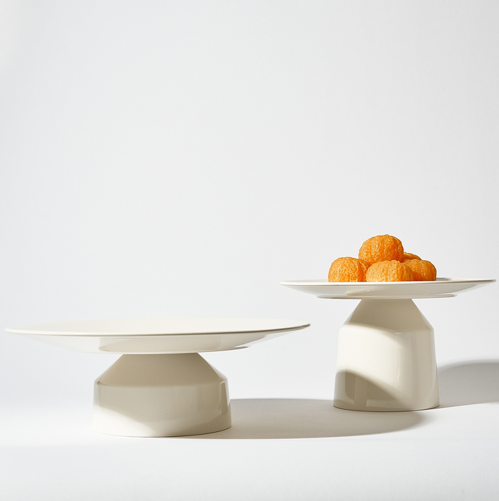 Two white plates, including a Solare Serving Tray on a stand, are displayed against a plain white background; the plate on the left is empty while the right one holds a small pile of orange round pastries or sweets.