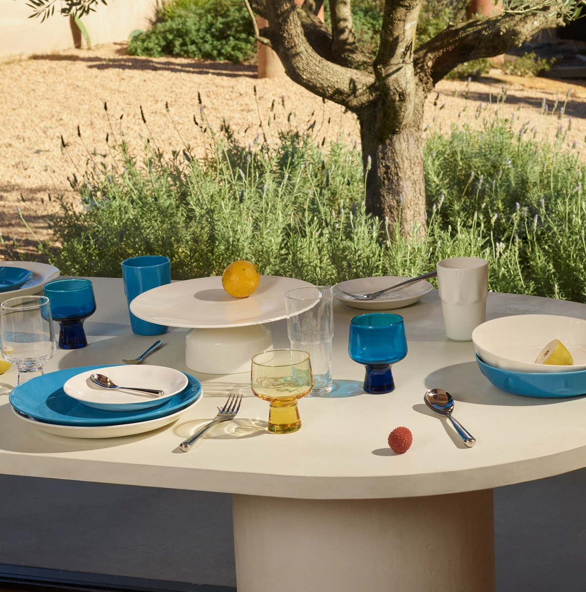 A white outdoor table is set with blue, white, and clear Iittala glassware and dishes. Three fruits are scattered beside the Solare Serving Tray. A tree and green bushes form the backdrop.