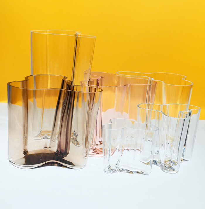 A pair of Alvar Aalto Vase Set of 2, featuring wavy-edged, translucent glass in clear and amber, is displayed on a white surface against a vibrant yellow background.