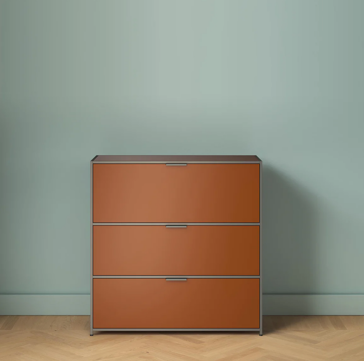 The Dita Secretaire Desk by Ligne Roset features orange-brown fronts and sleek handles, standing against a plain light blue wall on a wooden herringbone floor.