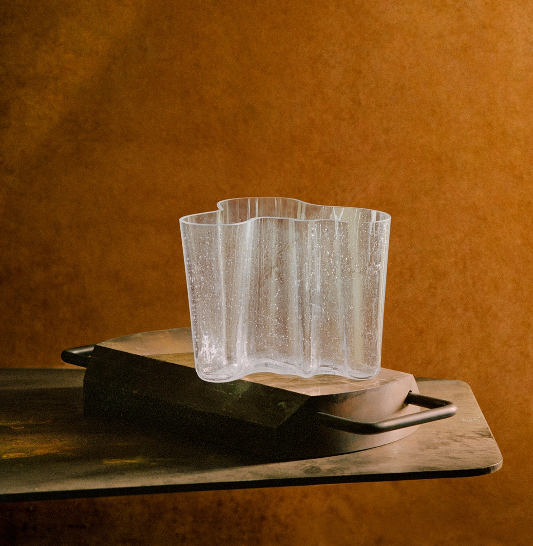 The Alvar Aalto Bubble Vase, with its clear wavy edges and water droplets, rests on a rectangular block on a dark metallic surface, set against a warm brown textured background.