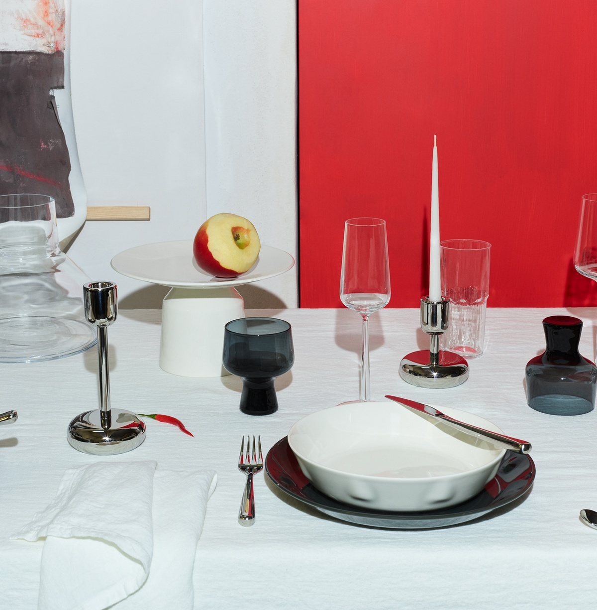 A modern table setting featuring Iittala glassware, white and black plates, silver cutlery, a white napkin, metal candle holders, a Solare Serving Tray with an apple and red chili pepper on a crisp white tablecloth. Red and white abstract backgrounds.