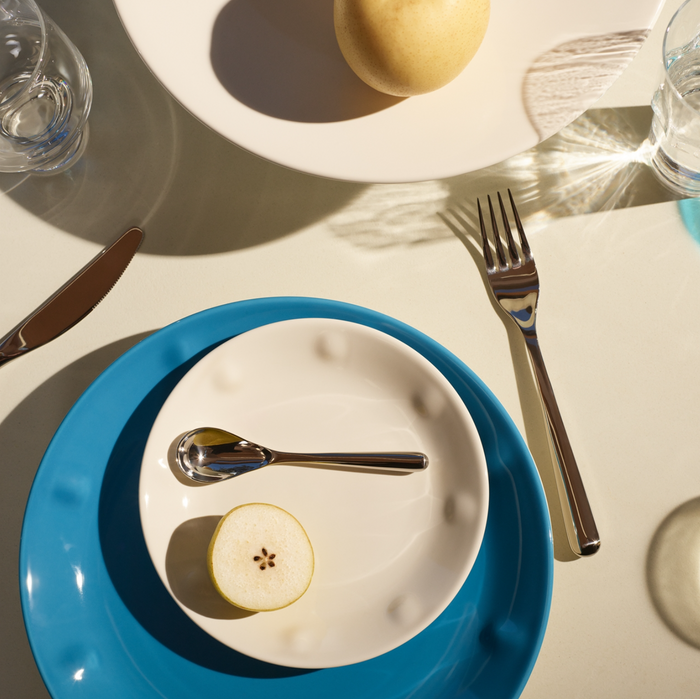 A table setting features a blue plate with a white plate, half-cut pear, and the Piano Cutlery Gift Box set (small spoon, fork, knife). Another white plate with a whole pear and glasses are visible in bright sunlight.