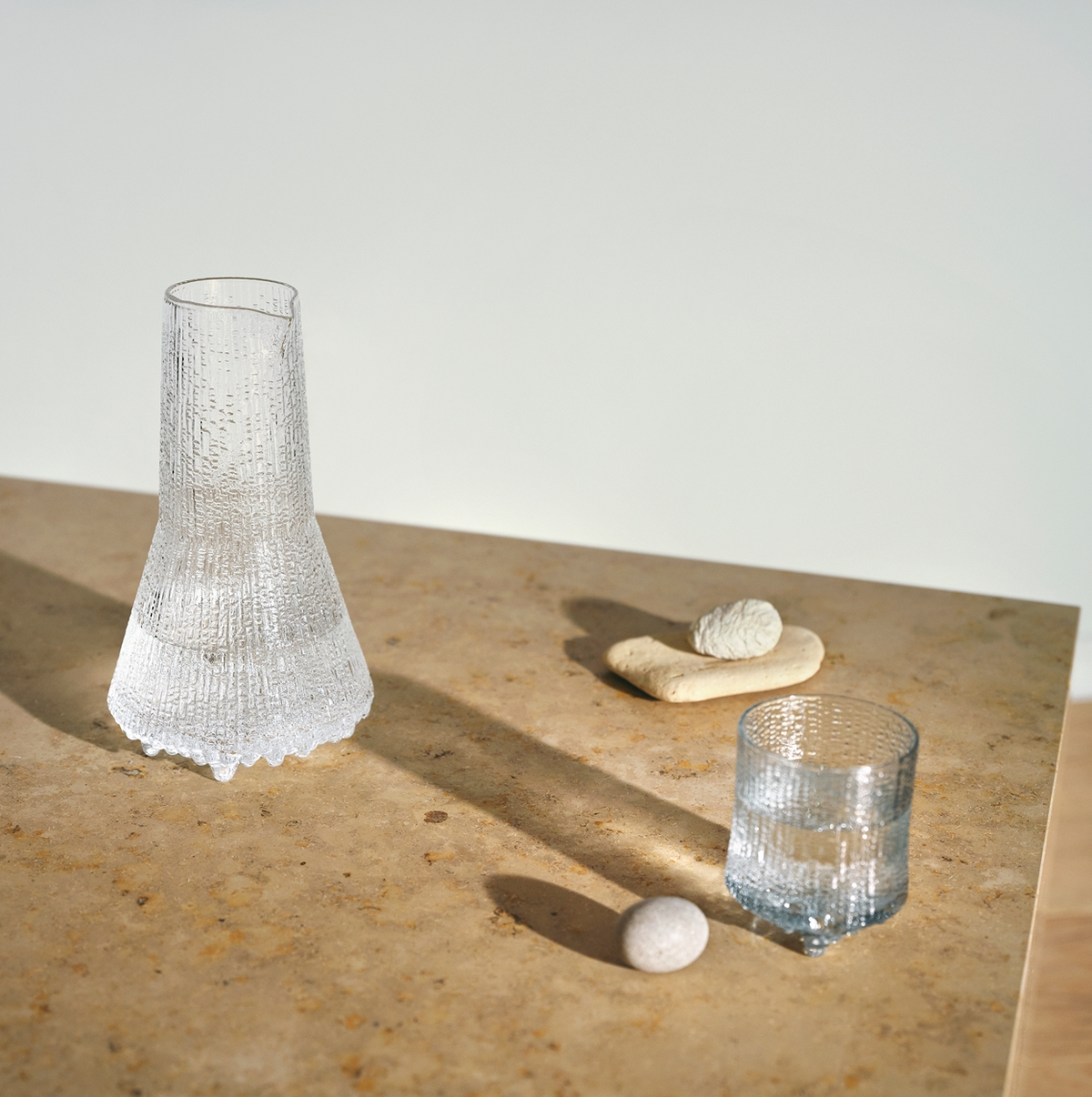 A clear Ultima Thule Pitcher 50cl sits on a light brown stone table with small stones and a beige decor piece, casting distinct shadows in soft sunlight—an ode to serene Nordic design.