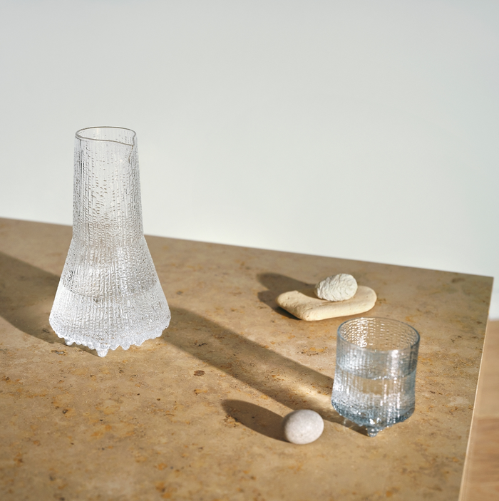 A clear Ultima Thule Pitcher 50cl sits on a light brown stone table with small stones and a beige decor piece, casting distinct shadows in soft sunlight—an ode to serene Nordic design.