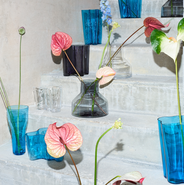 The Alvar Aalto Vase, 270 mm, in blue, clear, and dark tones holds pink and white anthuriums on concrete stairs against a light wall, highlighting striking Finnish design in a modern display.