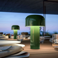 A Flos Bellhop Portable Lamp, IP54, in green glows stylishly on a wooden outdoor dining table set with plates and glasses, providing cordless illumination beside another lamp against a softly blurred evening backdrop.