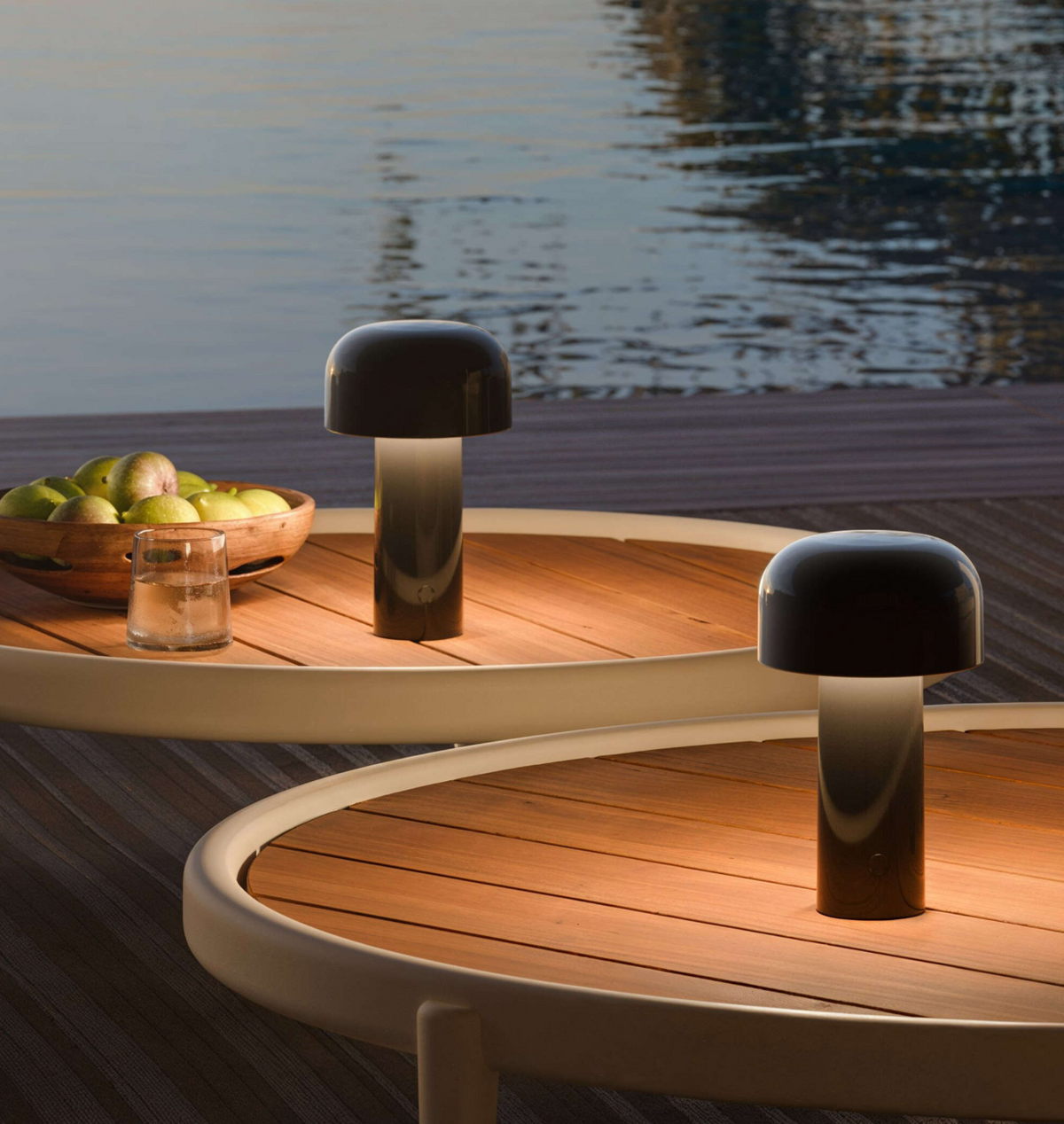 Two Flos Bellhop Portable Lamps (IP54) illuminate round wooden tables by the water; one table displays green apples and a glass tumbler, evoking a serene outdoor atmosphere at dusk.