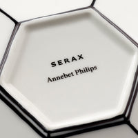 Close-up of a white ceramic hexagonal plate with delicate black edging, showcasing the Serax brand name and designer Annebet Philips’ signature in black lettering at the center. Its refined finish evokes the timeless elegance of fine bone china, making it a perfect pairing for your Serax Carte Blanche Cup and Saucer Set of 2.