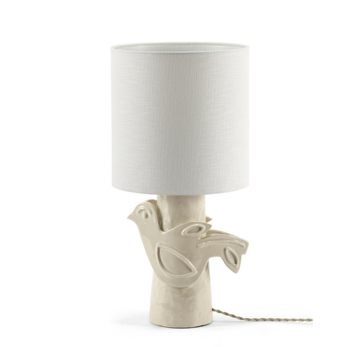 The Paloma Table Lamp in white by Serax is a handcrafted piece featuring a ceramic base shaped like a dove perched on a branch and topped with a cylindrical fabric shade; the power cord remains visible.