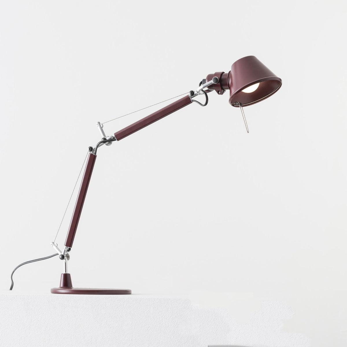 An Artemide Tolomeo Micro Desk Lamp in maroon, featuring a round base, angled adjustable arm, and illuminated bulb, is placed on a white surface with a plain white background.