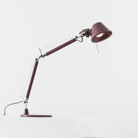 An Artemide Tolomeo Micro Desk Lamp in maroon, featuring a round base, angled adjustable arm, and illuminated bulb, is placed on a white surface with a plain white background.
