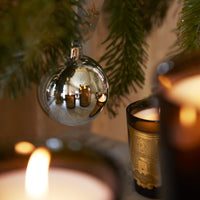 A luxury Trudon Holiday Candle 270g in a gold-labeled glass jar sits near a silver ornament hanging from a pine branch.