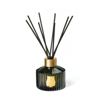 A black, ribbed glass Trudon Diffuser 350ml with a gold label and stopper features several thin black reed sticks fanned out from the top. This luxury home fragrance piece by Trudon elegantly scents any space while also serving as a sophisticated decor accent.