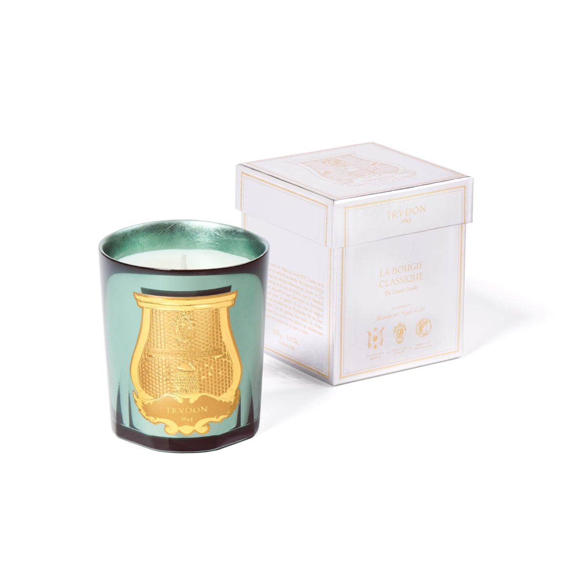 A green glass Trudon Holiday Candle 270g with a gold crest emblem next to its square white and gold box.