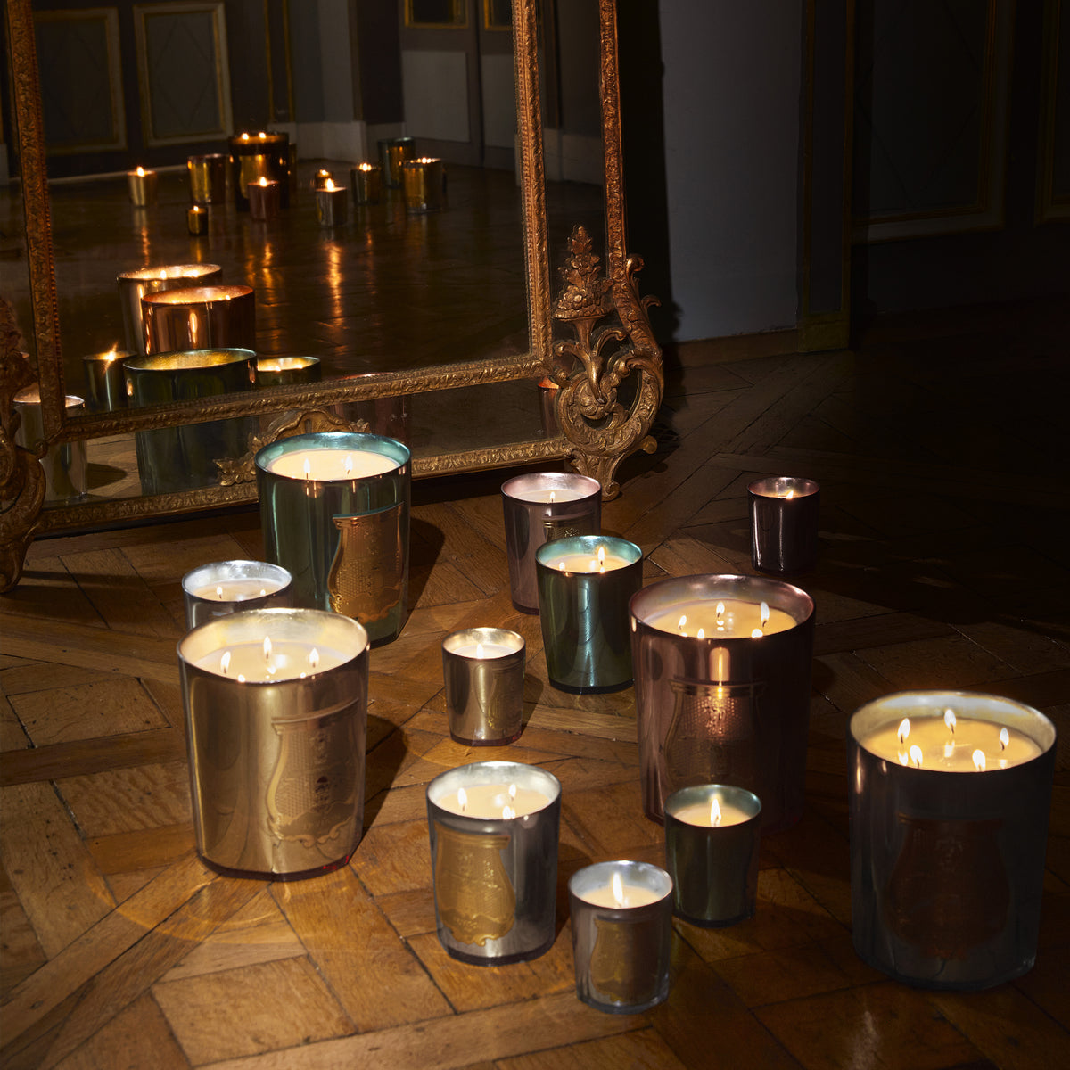 Many Trudon Holiday Candle 270g candles in metallic glass containers arranged on a wooden floor in front of an ornate floor mirror.