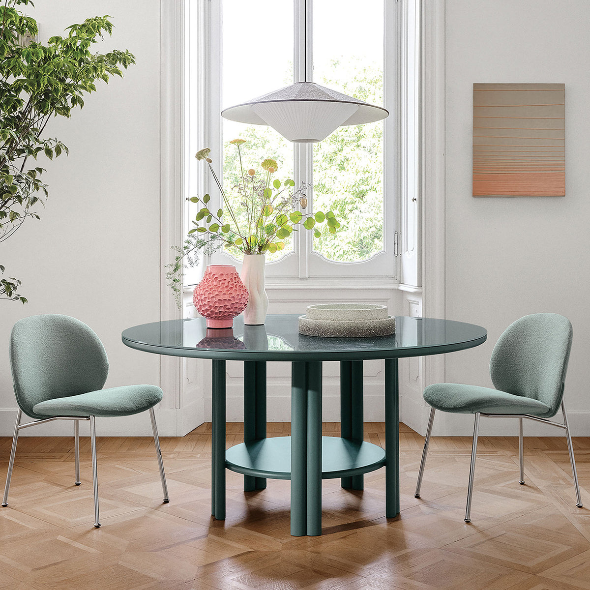 A modern dining area features the Saba Anam Dining Table, a round glass design, two light green upholstered chairs, a floral vase, pink decorative vase, and books. Large windows, art, and a leafy plant brighten the space.