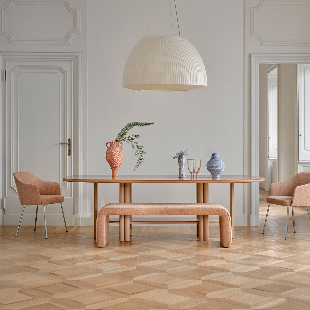 A modern dining room featuring the Saba Anam Dining Table, two beige chairs, a matching bench, decorative vases, and a large white pendant lamp, set against white paneled walls and light parquet flooring.
