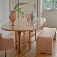 A modern dining room with the Saba Anam Dining Table in glossy beige, paired with a textured peach bench, matching chair, and decorative vases. Features light wood parquet floors, white walls, and a large window overlooking greenery.