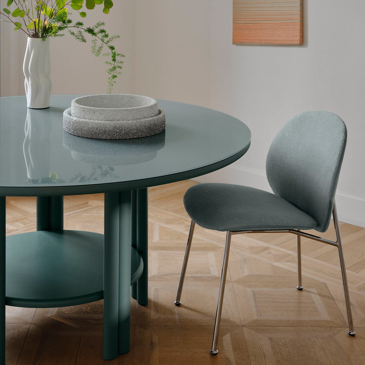 A modern dining area featuring Saba’s glossy green Anam Dining Table, a matching padded chair with metal legs, stacked stone bowls, a white vase with greenery on light wood parquet flooring, and framed art on the wall.