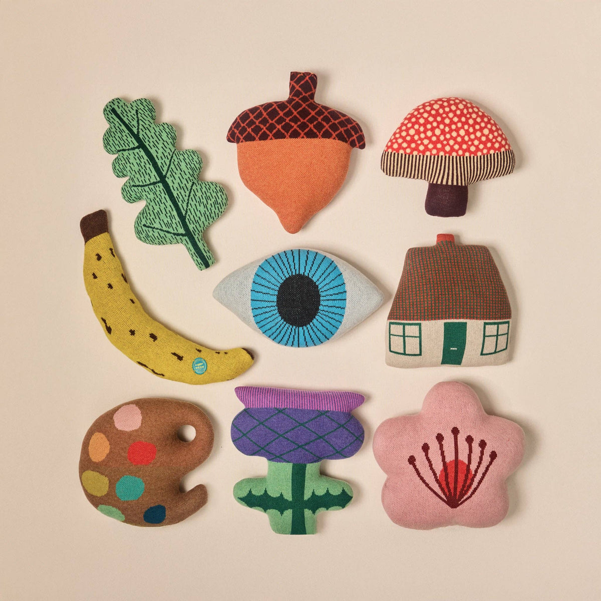 Donna Wilson’s Acorn Shaped Cushion is one of nine vibrant, soft fabric plush shapes—including an oak leaf, mushroom, banana, eye, house, artist palette, thistle flower, and cherry blossom—expertly knitted in Scotland from the finest materials and presented in a 3x3 grid against a neutral background.