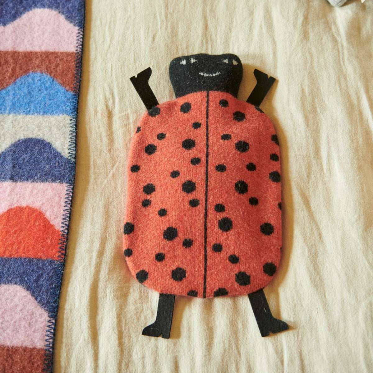 A Donna Wilson Ladybird Hot Water Bottle with a red body and black spots lies on a beige surface next to a colorful, wavy-patterned blanket.