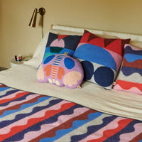 A bed is adorned with colorful, patterned pillows and a matching blanket featuring wavy stripes in red, blue, pink, and purple. At the center of the arrangement sits the Donna Wilson Totem Large Square Cushion - Red—an expertly crafted lambswool cushion made in the UK—while a cup rests on the bedside table.