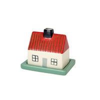 Introducing the Donna Wilson Little Hiding House by Donna Wilson—a small stoneware butter dish crafted in the shape of a house, complete with a red roof, white walls, black chimney, green door, and four charming windows. Set on a green rectangular base, this delightful piece features a hidden compartment that’s ideal for stashing tiny treasures or using as a keepsake box.