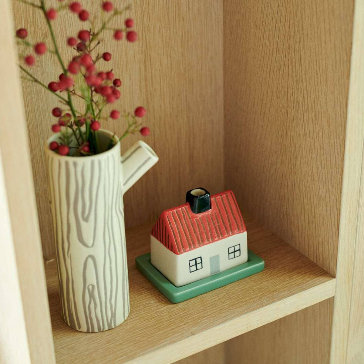 The Donna Wilson Little Hiding House, a charming stoneware piece featuring a red roof and a secret compartment for storing tiny treasures, sits beside a tall vase with a wood pattern and red berries, both beautifully displayed on a light wooden shelf.