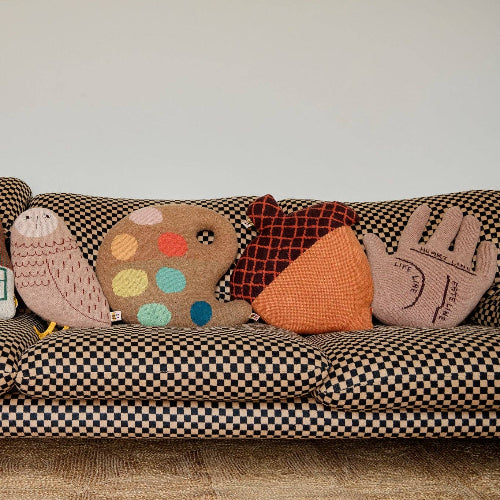 A checkered sofa adorned with five playful decorative pillows, including the Donna Wilson Acorn Shaped Cushion—each handcrafted from soft lambswool for comfort and charm—which also features two hand-shaped cushions, a house-shaped pillow, a polka dot design, and a bird-shaped cushion.