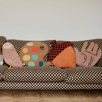 A checkered sofa adorned with five playful decorative pillows, including the Donna Wilson Acorn Shaped Cushion—each handcrafted from soft lambswool for comfort and charm—which also features two hand-shaped cushions, a house-shaped pillow, a polka dot design, and a bird-shaped cushion.