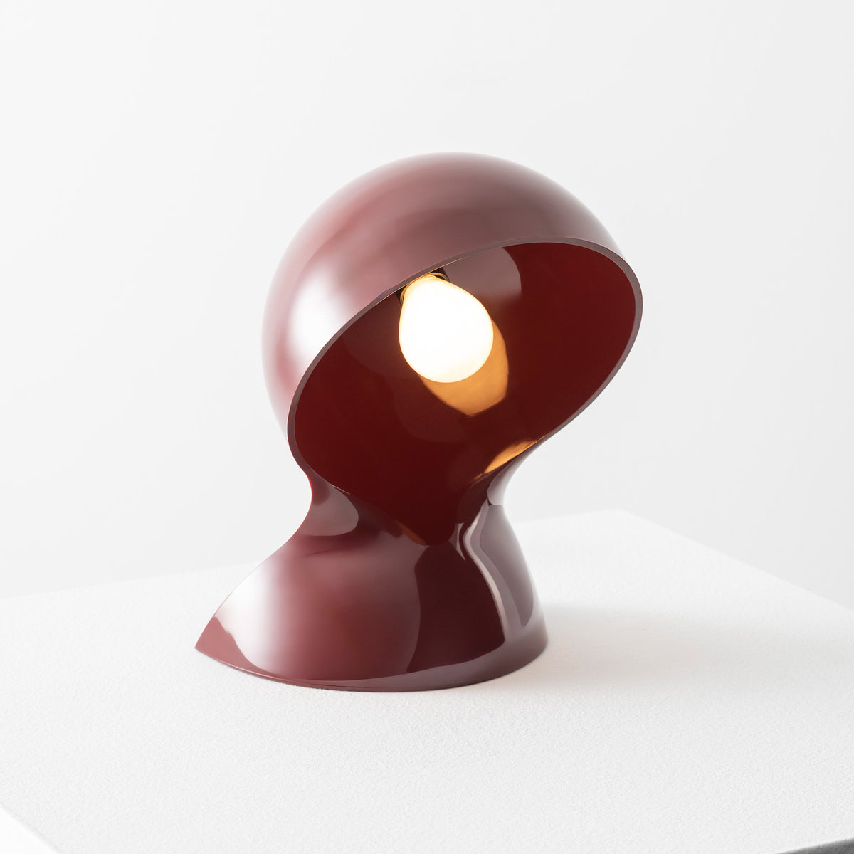 A sculptural maroon Dalu Table Light by Artemide, featuring a glossy finish and a distinctive curved, helmet-like form that partially encases a glowing bulb, sits on a white surface against a plain background.