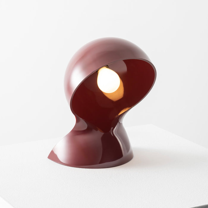A sculptural maroon Dalu Table Light by Artemide, featuring a glossy finish and a distinctive curved, helmet-like form that partially encases a glowing bulb, sits on a white surface against a plain background.
