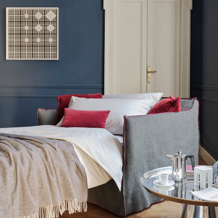 A cozy bedroom with a Gervasoni Ex-Display Ghost Single Sofa Bed 11 in gray with removable cover, accented by white and pink pillows, a beige blanket, blue walls, abstract art, and a round glass table set with coffee pot and cups.