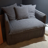A gray Gervasoni Ex-Display Ghost Single Sofa Bed 11 with three matching cushions and a removable cover sits against a dark wall on a light concrete floor.