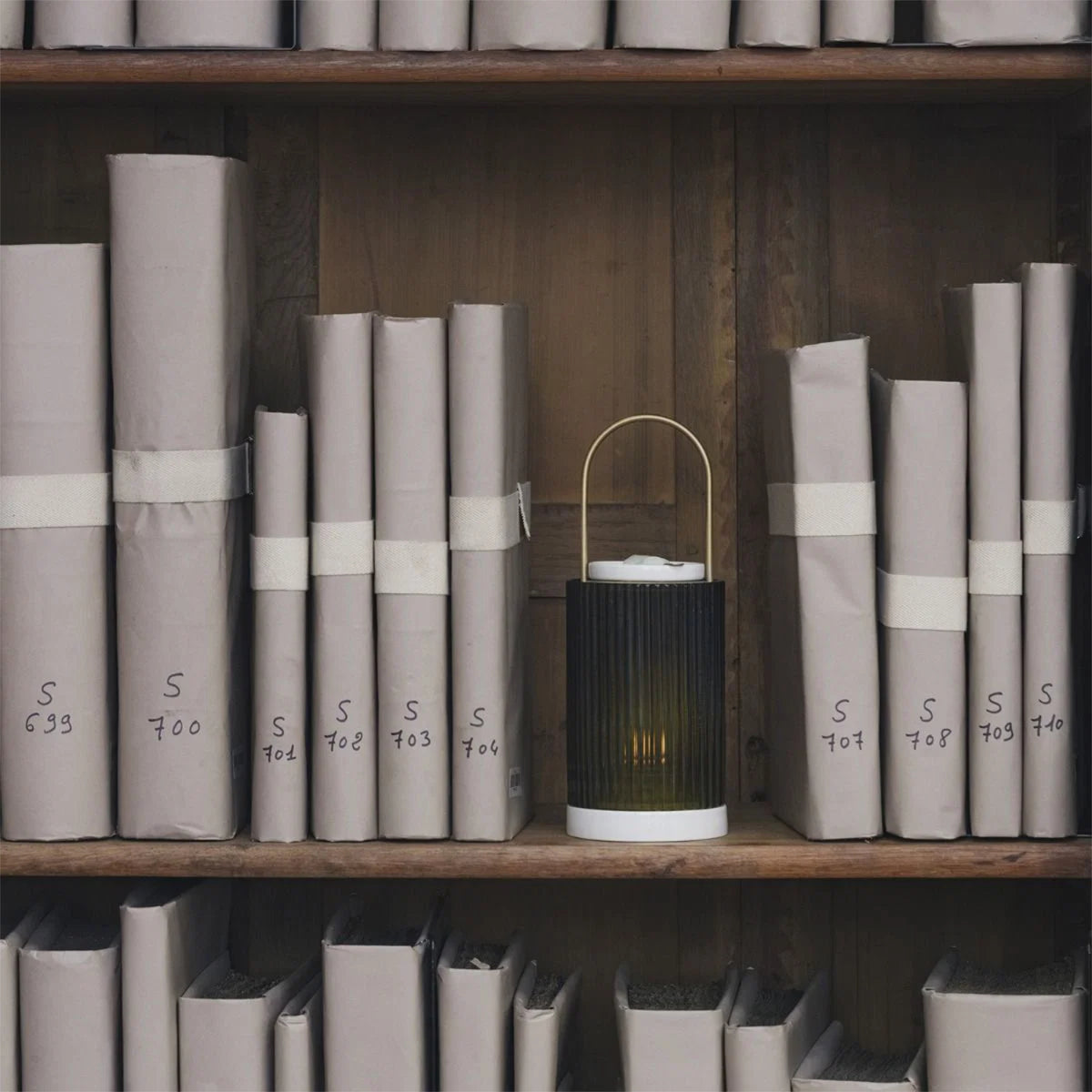 A cylindrical, ribbed Trudon La Promeneuse candle warmer with a brass handle sits on a wooden shelf among numbered, paper-wrapped books.