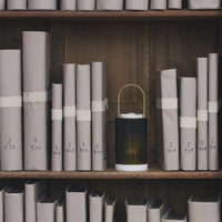 A cylindrical, ribbed Trudon La Promeneuse candle warmer with a brass handle sits on a wooden shelf among numbered, paper-wrapped books.