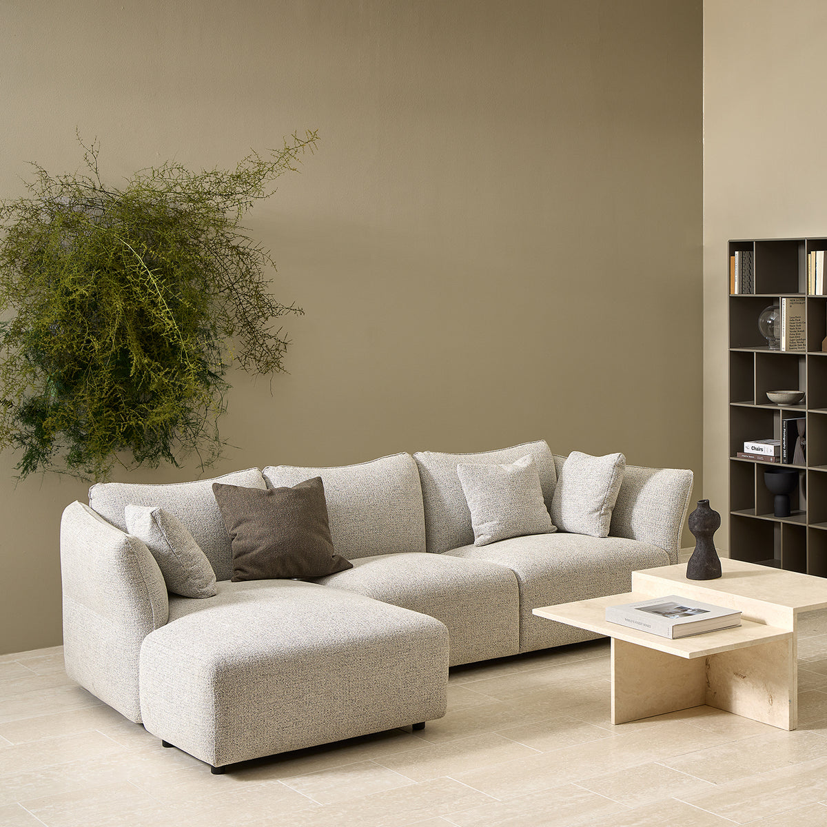 A modern living room with the Flexlux Ex-Display Melfi 3-Seater Sofa in Veneto Nature, neutral pillows, a beige square coffee table with a sculpture, wall-mounted plant, and a gray shelf with books and decor against a taupe wall.