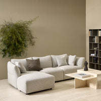A modern living room with the Flexlux Ex-Display Melfi 3-Seater Sofa in Veneto Nature, neutral pillows, a beige square coffee table with a sculpture, wall-mounted plant, and a gray shelf with books and decor against a taupe wall.