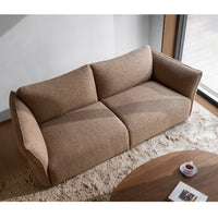 A Flexlux Ex-Display Melfi 3-Seater Sofa in Veneto Nature stands on a beige rug beside a round wooden coffee table with a cup and saucer, near a large window in a modern, well-lit living room.