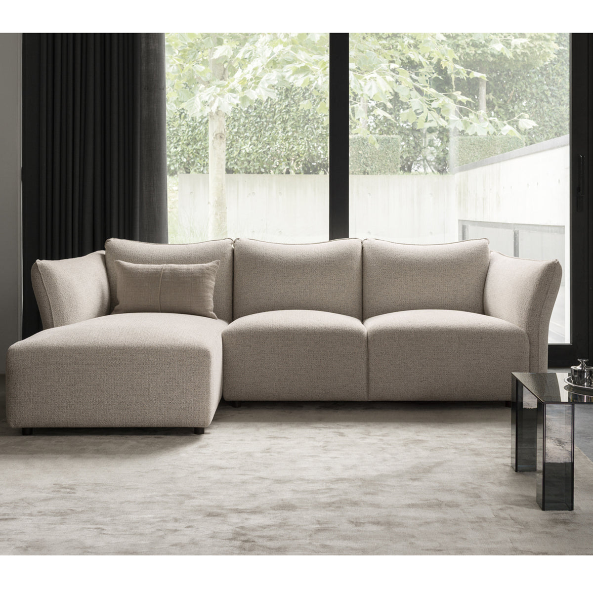 The Ex-Display Melfi 3-Seater Sofa in Veneto Nature by Flexlux sits on a light carpet near tall windows with greenery outside and a glass coffee table—ideal for a modern lounge aesthetic.