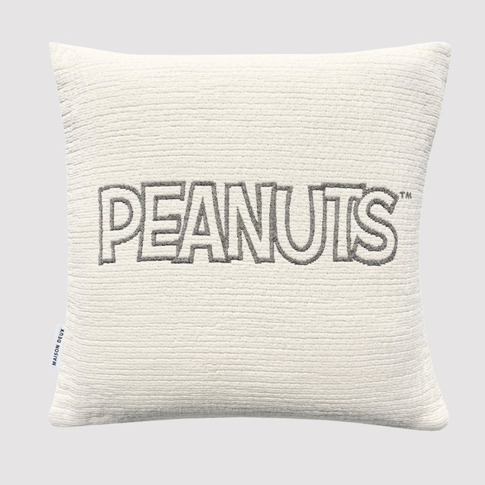 The Peanuts Cushion is a square, white cotton cushion with a textured surface. "PEANUTS" is embroidered in bold gray letters across the center, and a small side tag decorates the left edge, taking inspiration from Maison Deux’s Snoopy cushion.