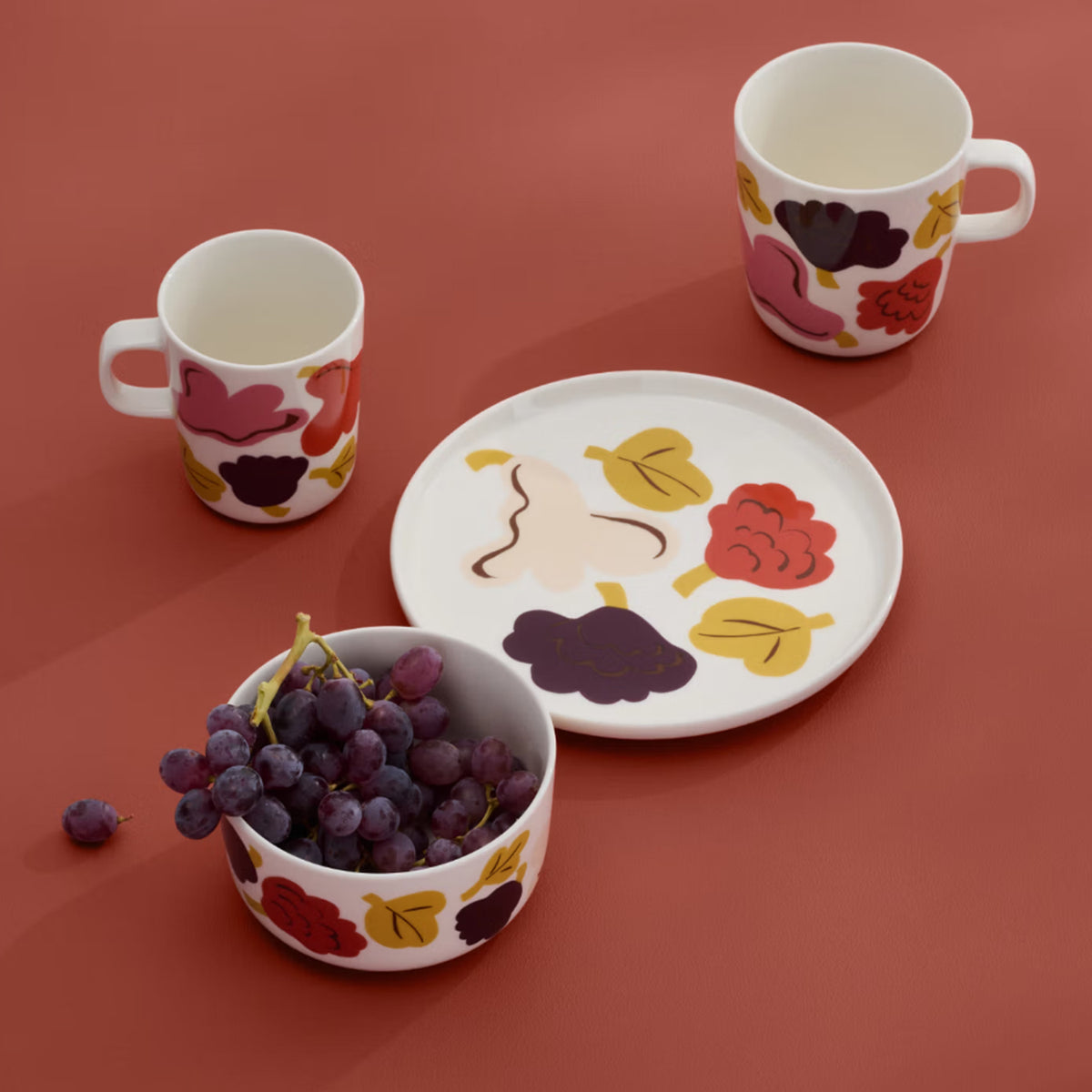 A white Marimekko Kukasta Kukkaan Bowl (4dl) featuring colorful abstract fruit and leaf patterns sits on a red surface, filled with grapes. Two matching mugs and an Oiva plate with similar vibrant designs accompany the bowl.