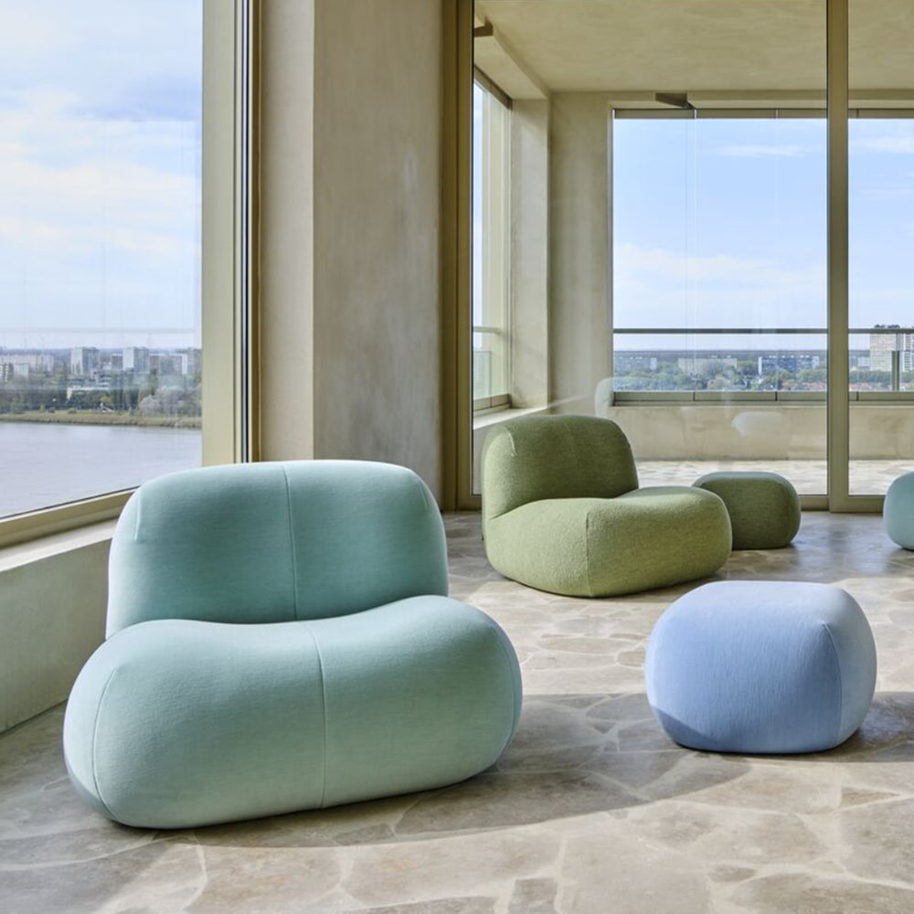 Softly contoured pastel chairs and a blue-green Pukka Small Ottoman by Ligne Roset (Kvadrat Gentle 2) complement a bright, modern living space with large windows, stone floors, and views of the city and river.