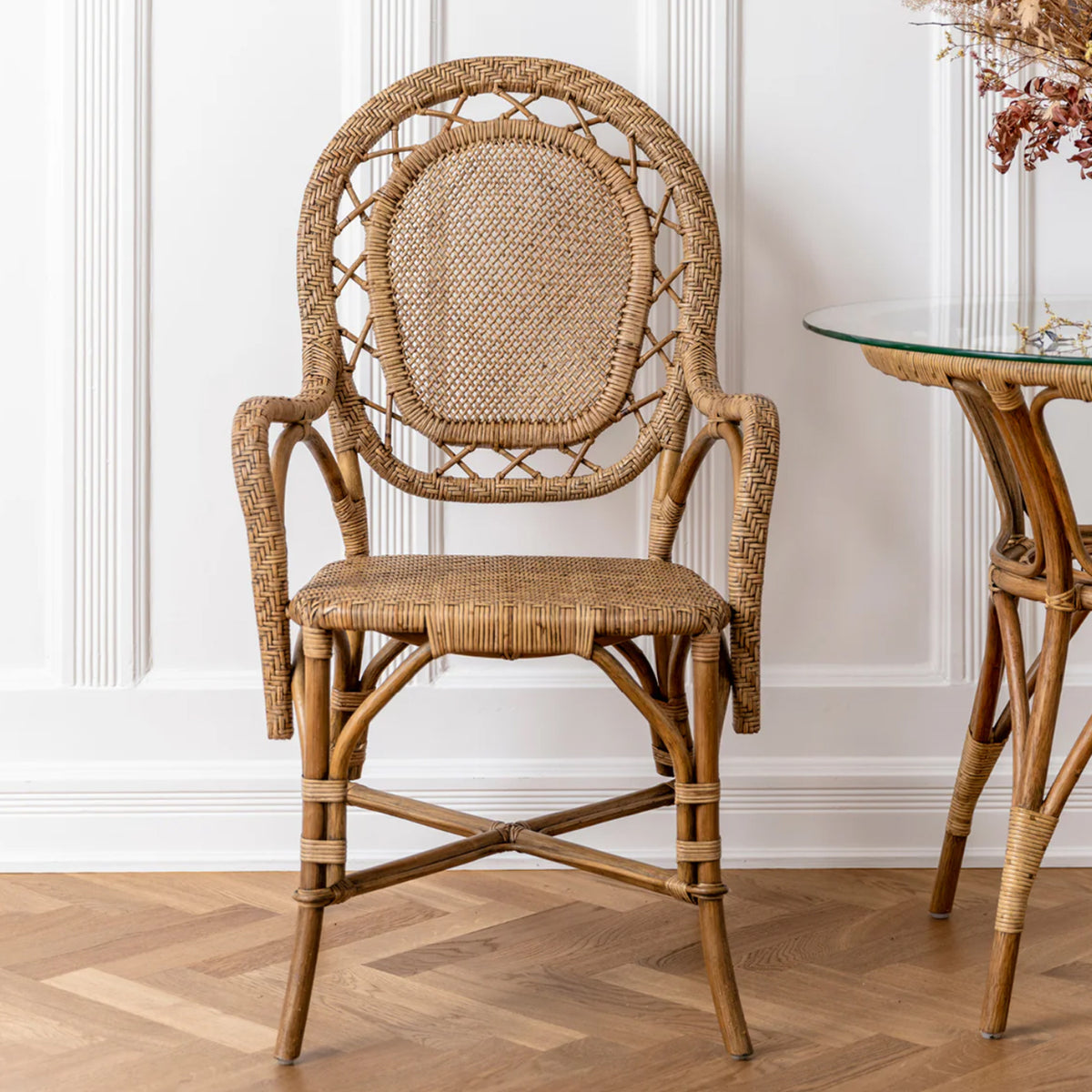 The Romantica Wicker Chair by Sika Design, featuring a rounded back and intricate woven patterns, stands on a wooden floor beside a glass-topped side table with a rattan base in a white-paneled room.