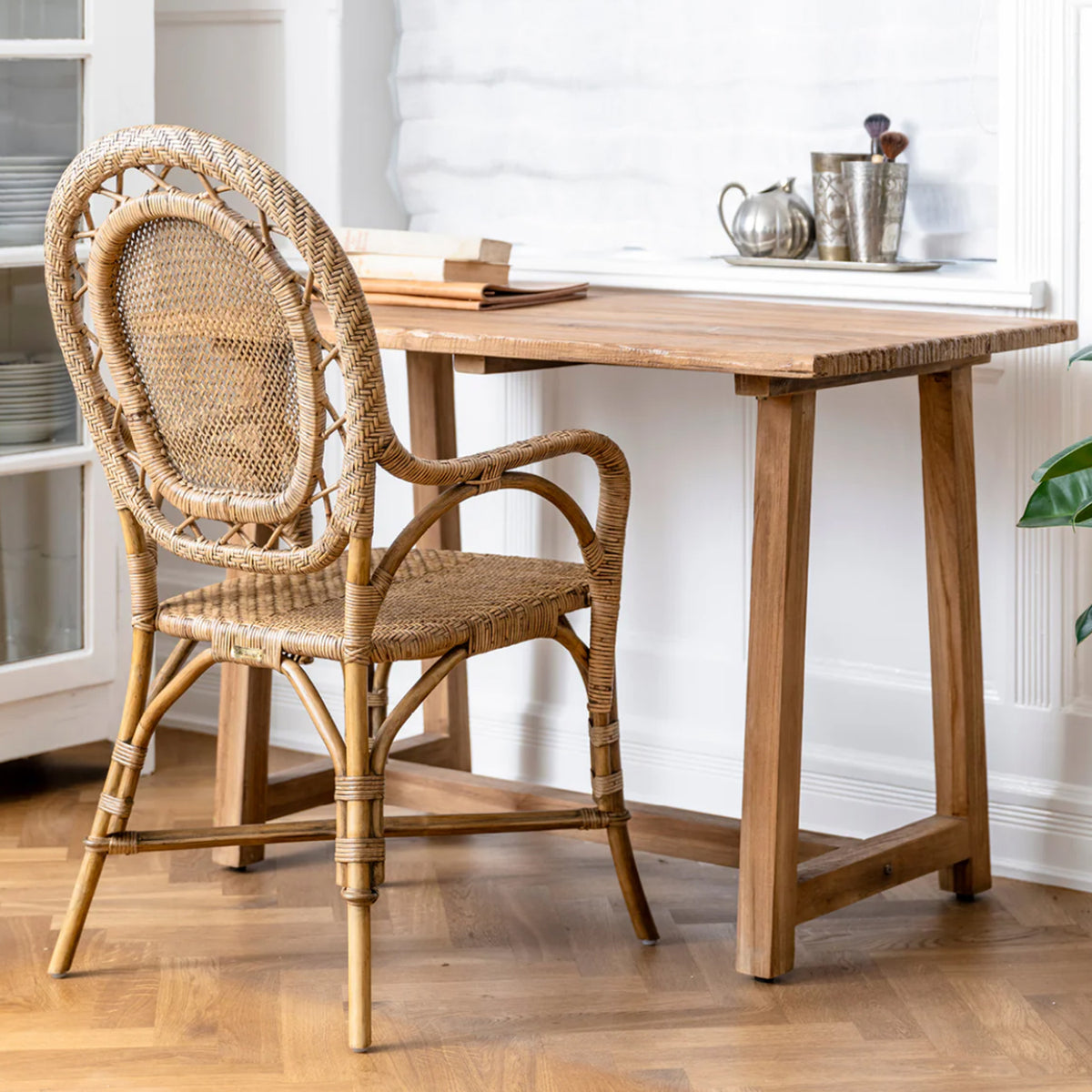 A bright room features the Romantica Wicker Chair by Sika Design at a wooden desk with books and a teapot set. The chair’s woven back complements the wood floor, white walls, and a nearby plant.
