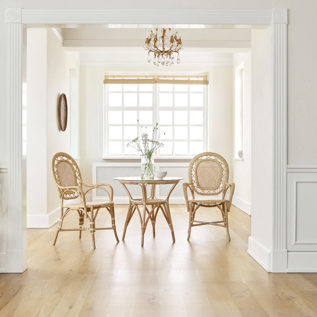 A bright dining nook features a glass round table and three Sika Design Romantica Wicker Chairs on light wood floors. Natural light streams through the window blinds, illuminating a small vase of flowers on the table.