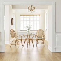A bright dining nook features a glass round table and three Sika Design Romantica Wicker Chairs on light wood floors. Natural light streams through the window blinds, illuminating a small vase of flowers on the table.