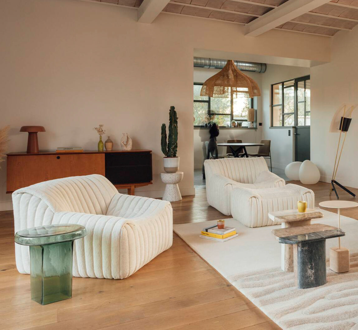 A cozy living room with two cream-colored Ligne Roset Sandra Armchairs, quilted upholstery, a glass side table, neutral rug, wood floors, mid-century console, modern decor, and a kitchen view add charm.